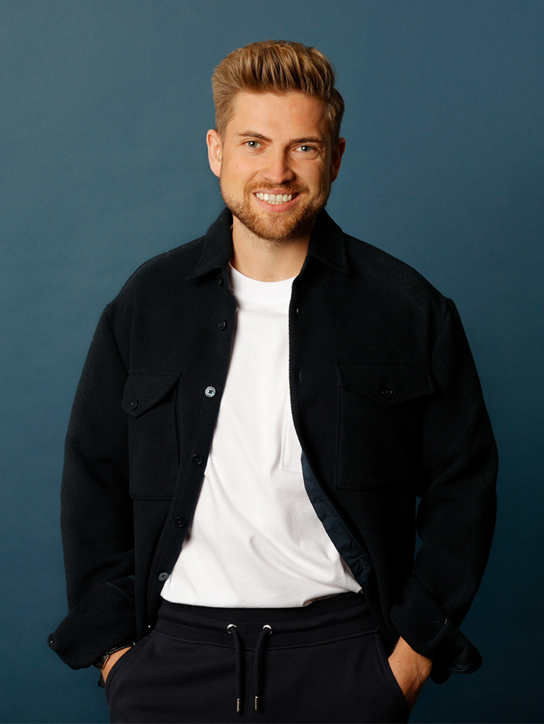 Ein lächelnder Mann mit blonden Haaren, weißem T-Shirt und schwarzer Jacke, vor blauem Hintergrund.