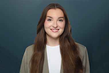 Eine lächelnde junge Frau mit langen braunen Haaren vor einem dunkelblauen Hintergrund.