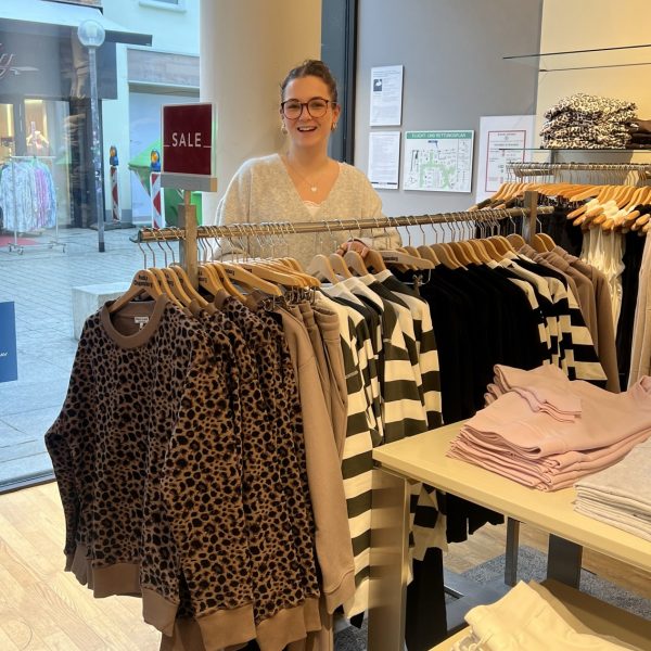 Eine Person steht lächelnd hinter einem Kleiderständer in einem Bekleidungsgeschäft. Auf dem Ständer hängen verschiedene Kleidungsstücke, darunter gestreifte und leopardengemusterte Oberteile. Ein Schild mit der Aufschrift 'Sale' ist sichtbar. Im Hintergrund ist ein Schaufenster mit Straßenansicht zu sehen.