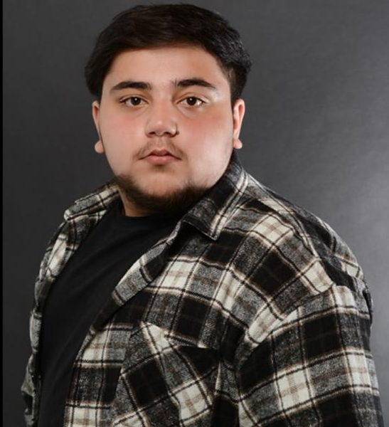 Ein junger Mann mit dunklen Haaren und Bart trägt ein kariertes Hemd und ein schwarzes T-Shirt vor einem grauen Hintergrund.