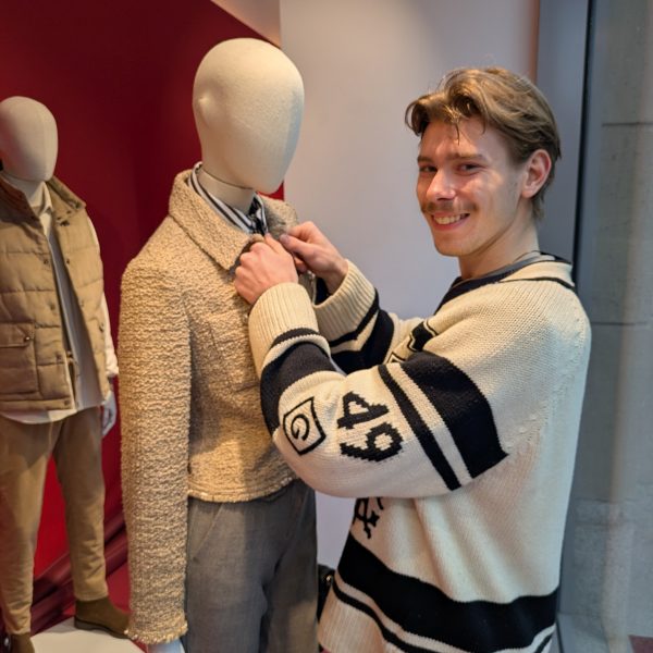 Ein lächelnder Mann in einem cremefarbenen Pullover mit schwarzen Streifen richtet eine Krawatte an einer Schaufensterpuppe in einem Ausstellungsraum, die mit einem beigen Jackett und grauer Hose bekleidet ist.