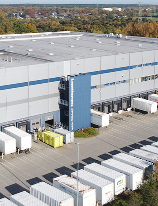 Luftaufnahme eines großen Lagerhauses mit mehreren Laderampen und geparkten Anhängern auf dem Parkplatz vor dem Gebäude.