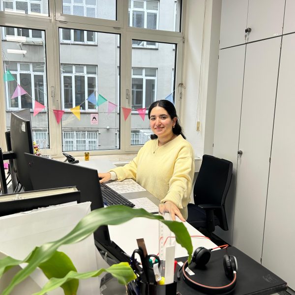 Eine Frau in einem gelben Pullover sitzt lächelnd an einem Schreibtisch in einem Büro und arbeitet am Computer. Im Hintergrund sind bunte Wimpel vor einem großen Fenster sichtbar.